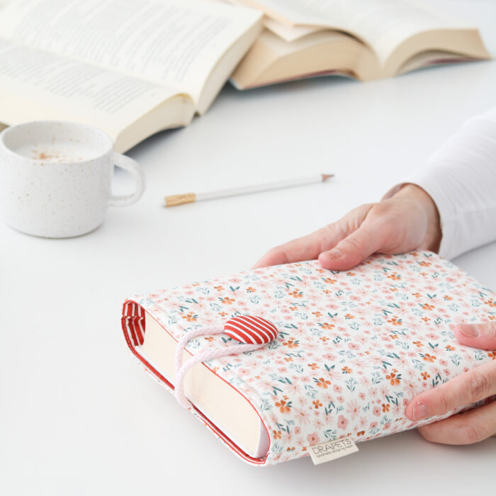 Funda de Libro confeccionada con tela estilo romántico de flores en rosa, toques verdes y teja combinada con un forro interior de rayas en blanco y teja. Funda acolchada para proteger tus lecturas.